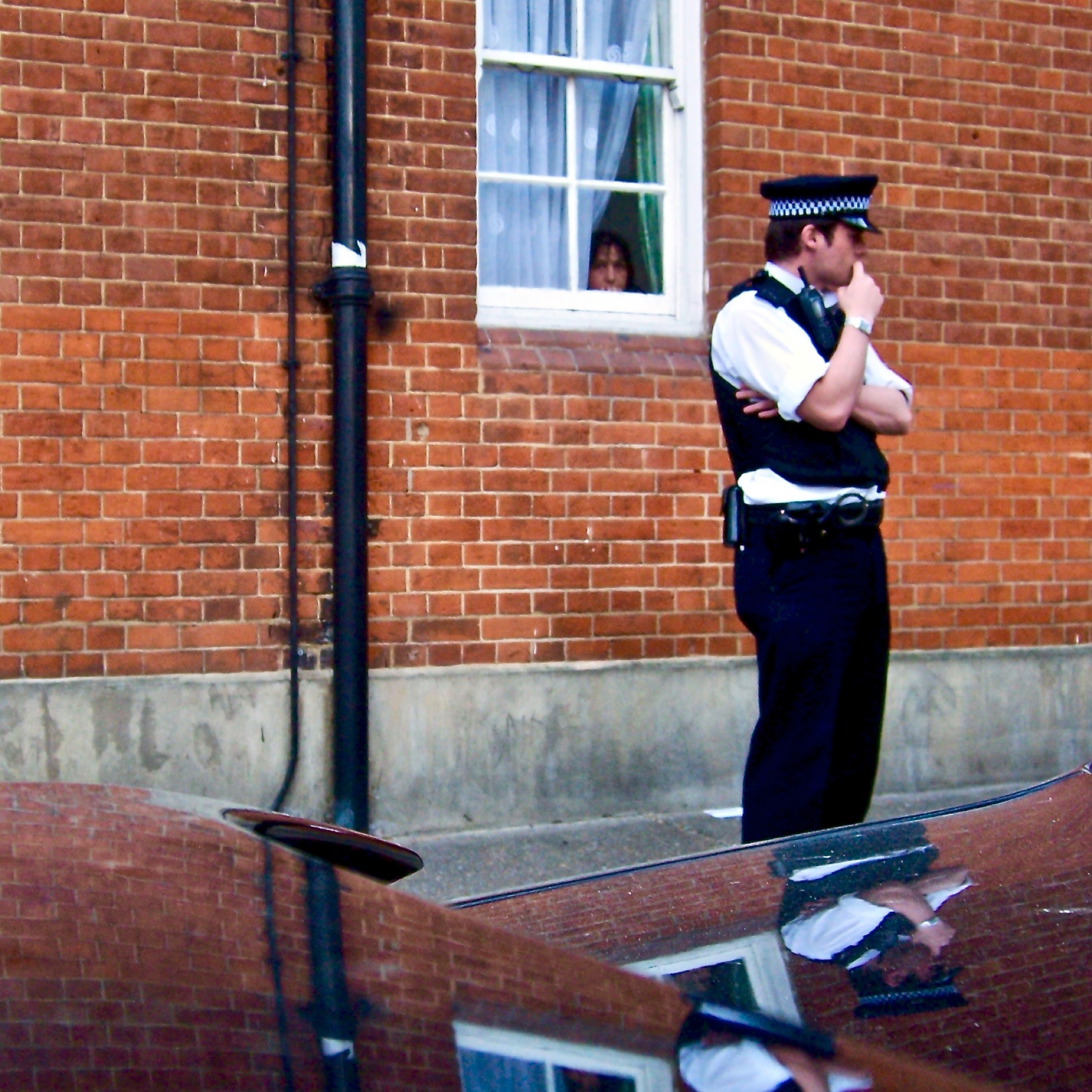 A pensive police officer stands guard outside a brick building, while a curious figure peeks through a window curtain.