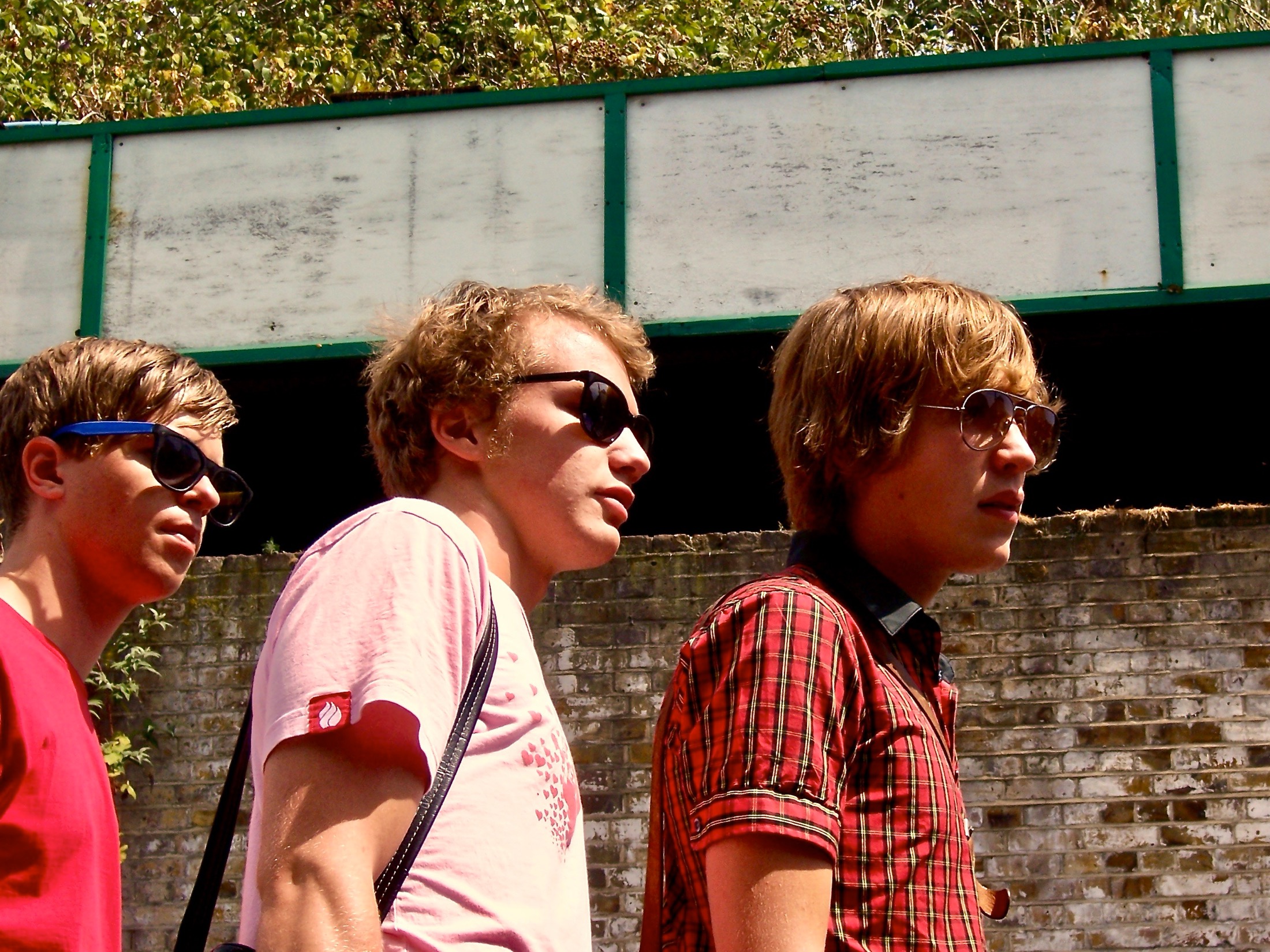 Three young men walk side by side in sunglasses, exuding a cool vibe against a weathered urban backdrop.