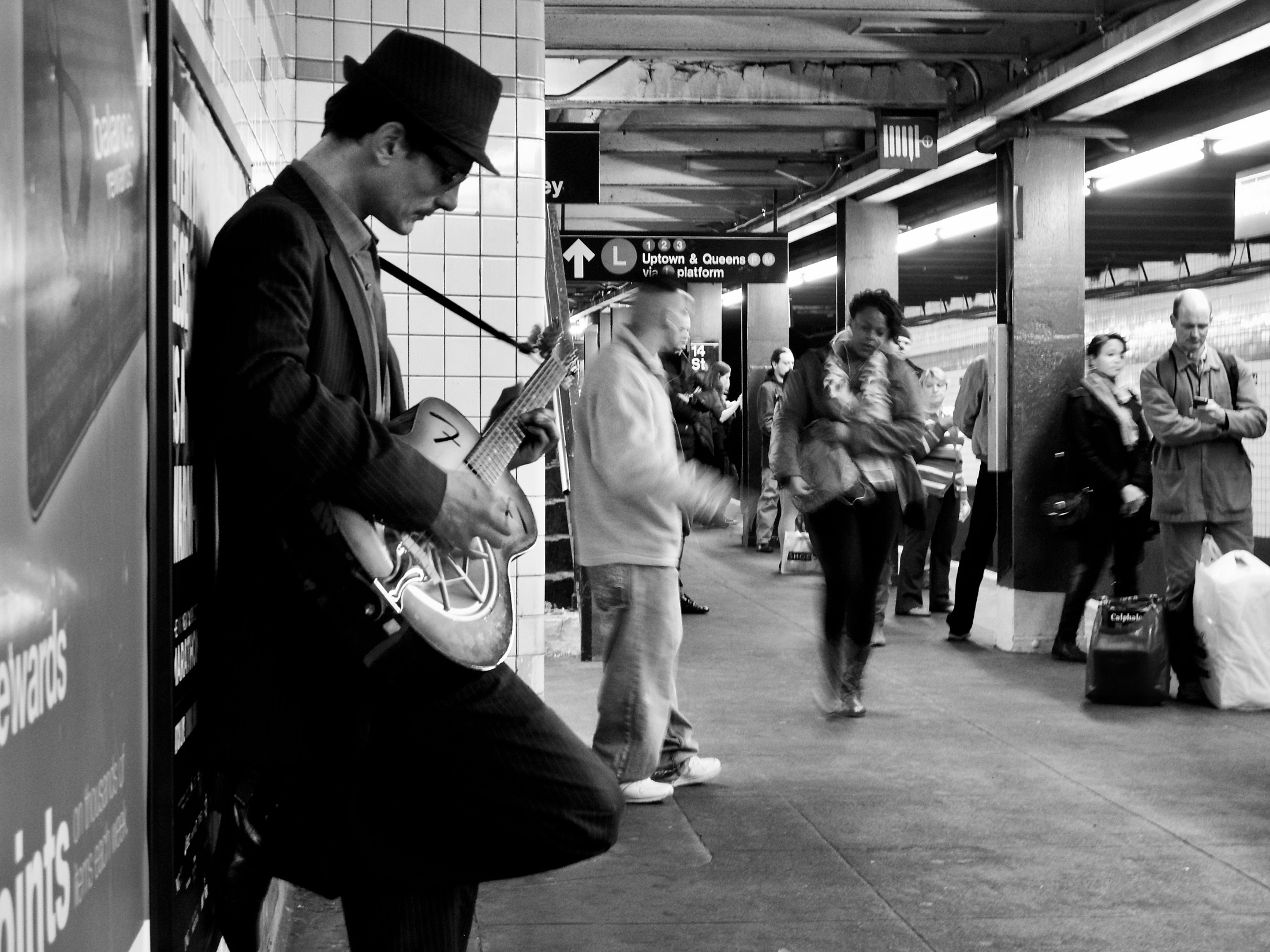A musician in a fedora strums his guitar against a subway wall, surrounded by hurried commuters in the bustling station.