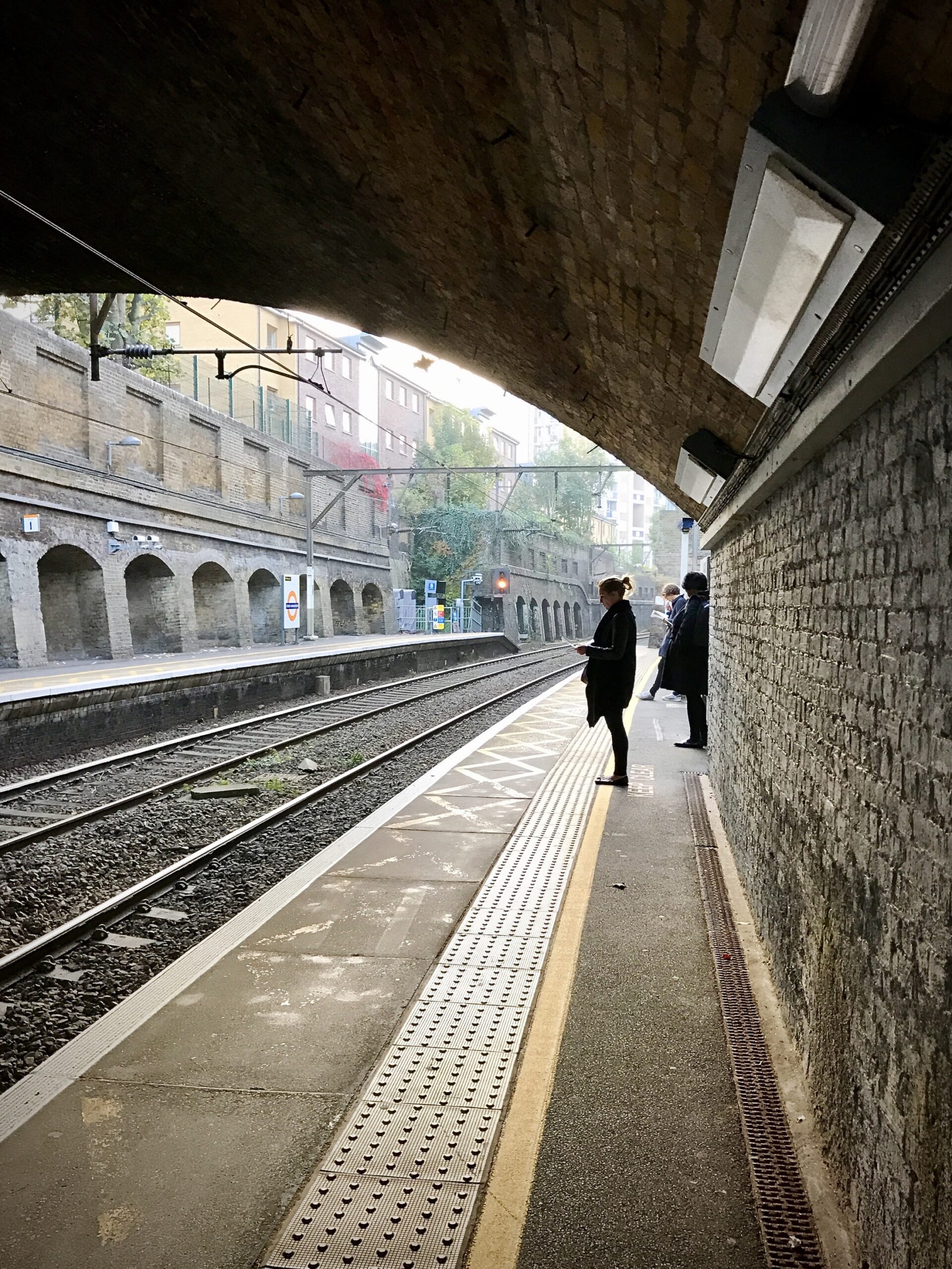 Passengers wait quietly on a dimly lit train platform, framed by an arching brick overpass and scattered autumn leaves.