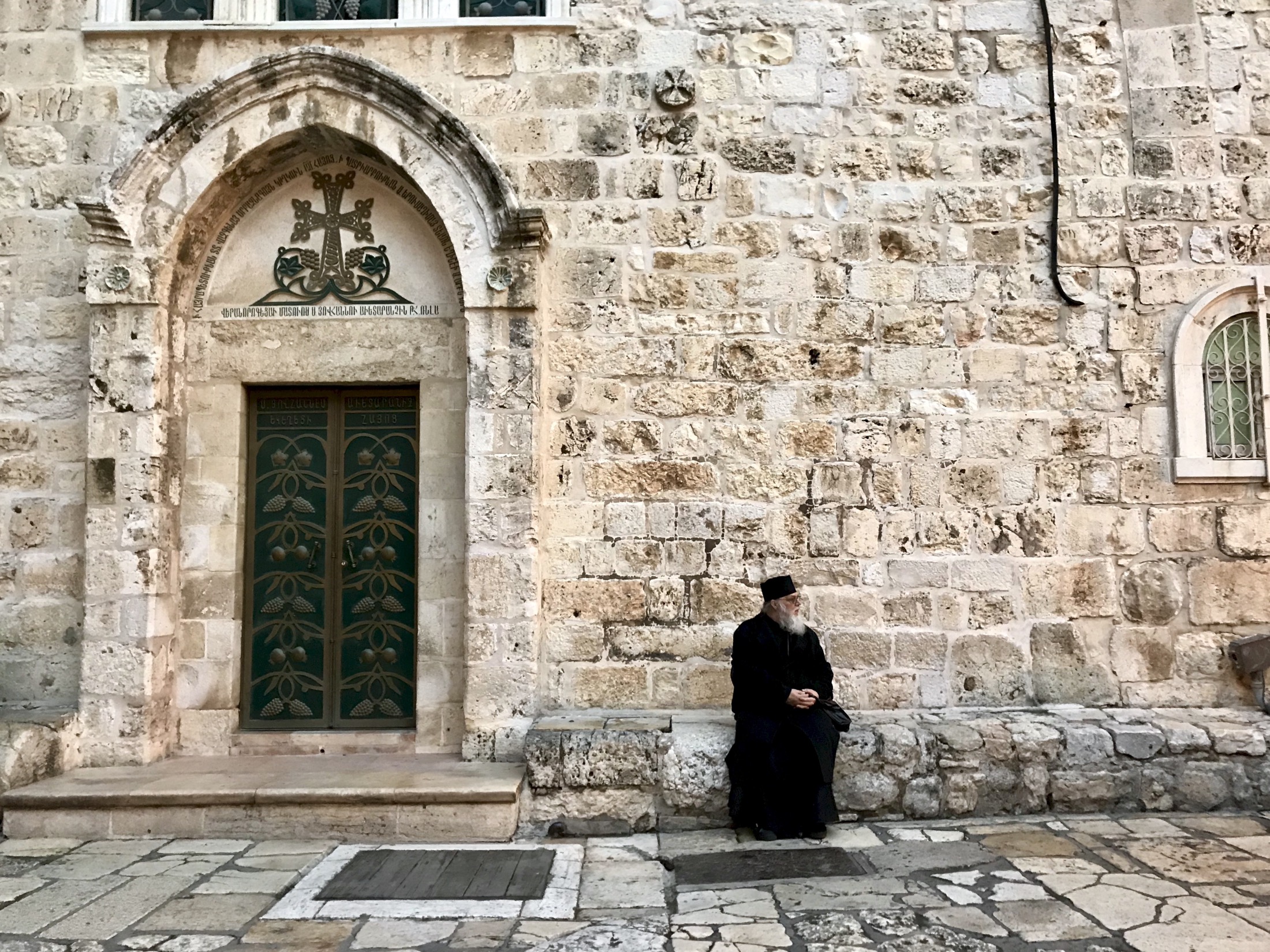 A bearded man in dark robes sits pensively against a rustic stone wall, absorbed in the serene atmosphere of the ancient setting.