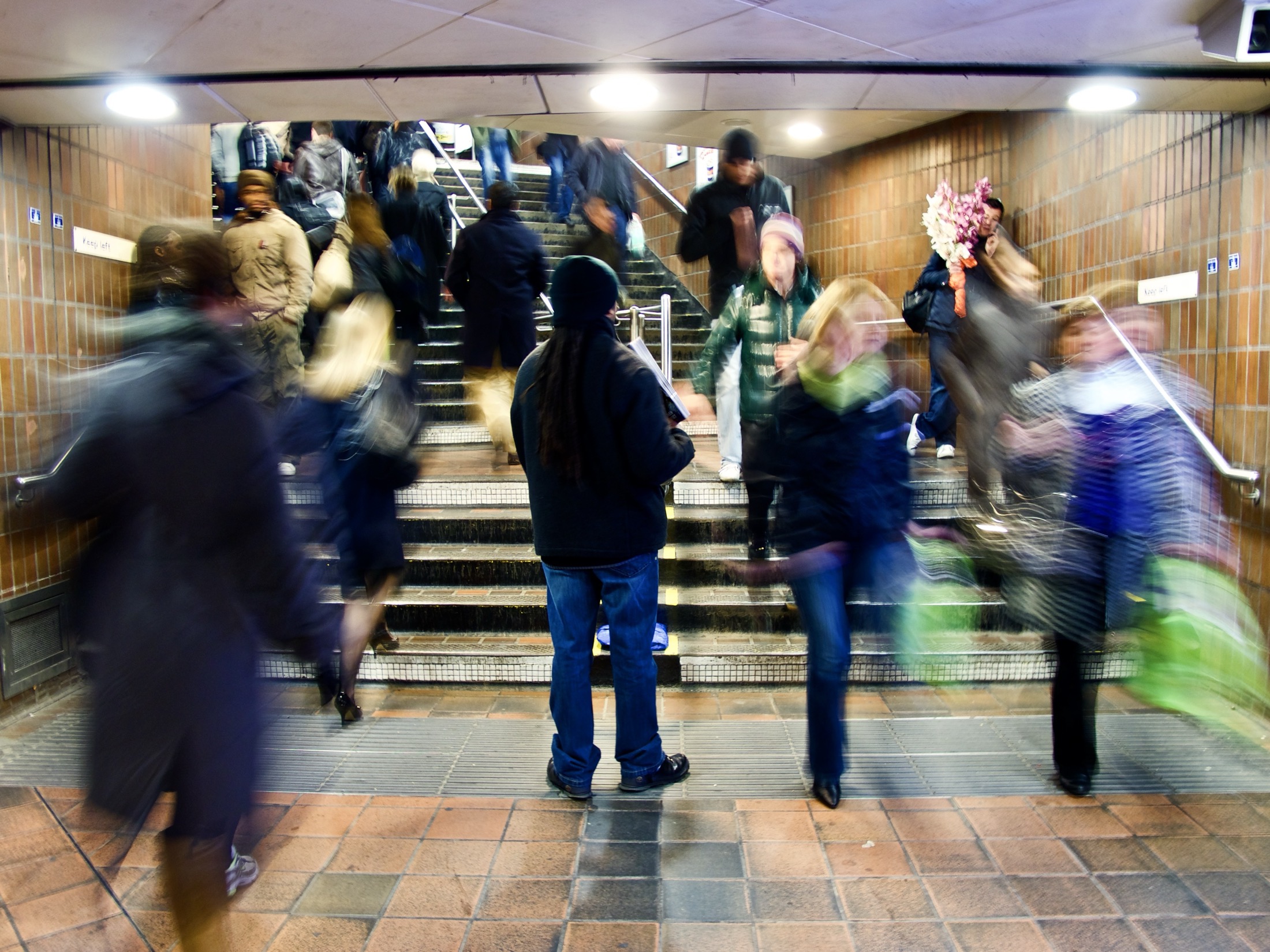 A single man is standing in a bustling subway entrance filled with blurred commuters rushing up and down the stairs, creating a lively urban scene.