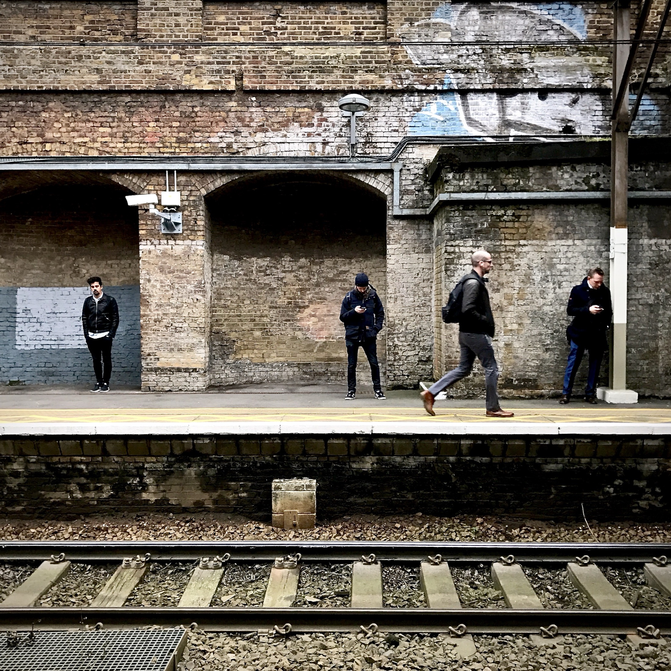 Four solitary figures stand at a train platform, immersed in their own worlds against a backdrop of faded brick and graffiti.