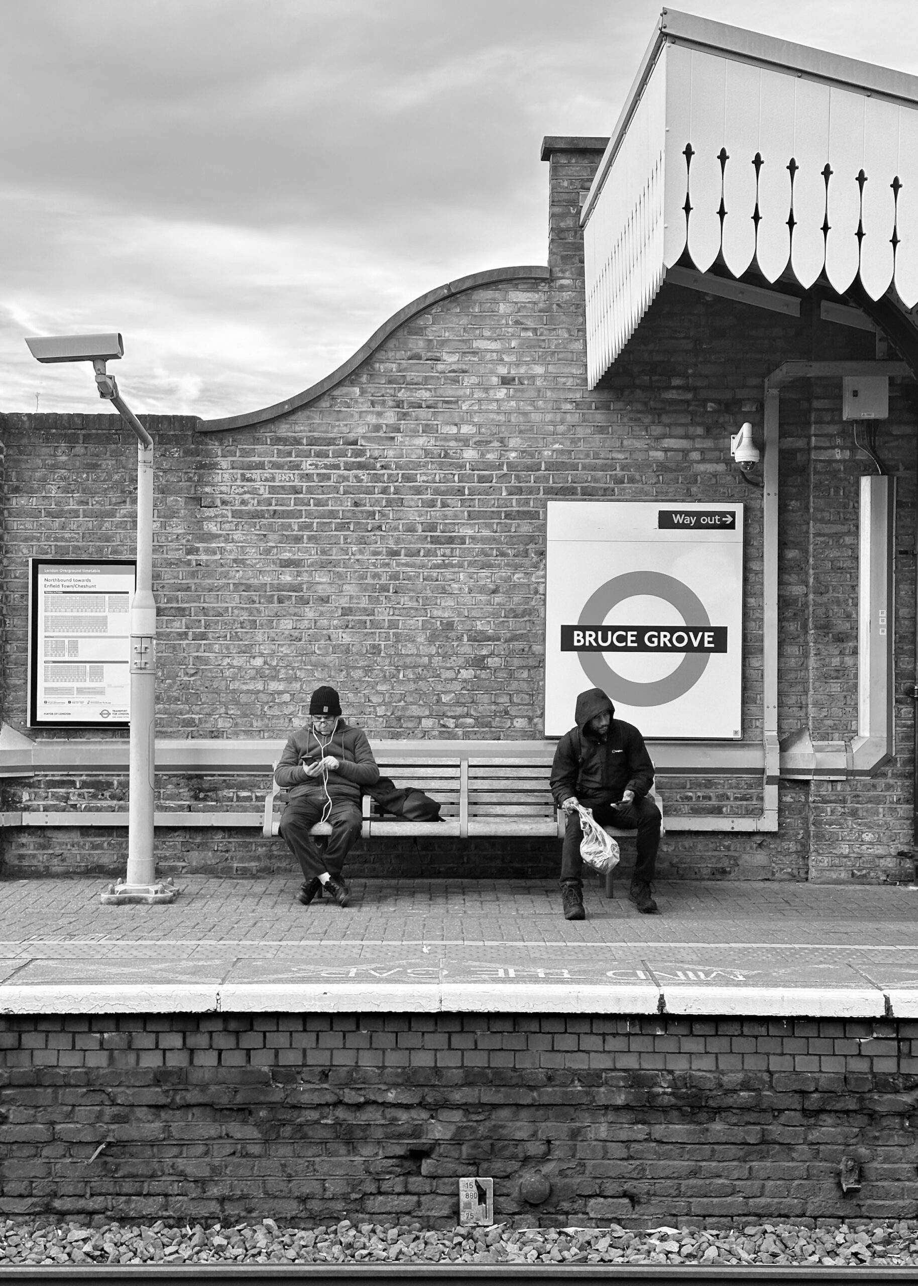 Two men sit quietly on a platform bench, immersed in their thoughts as the gray sky looms above the lonely brick station.