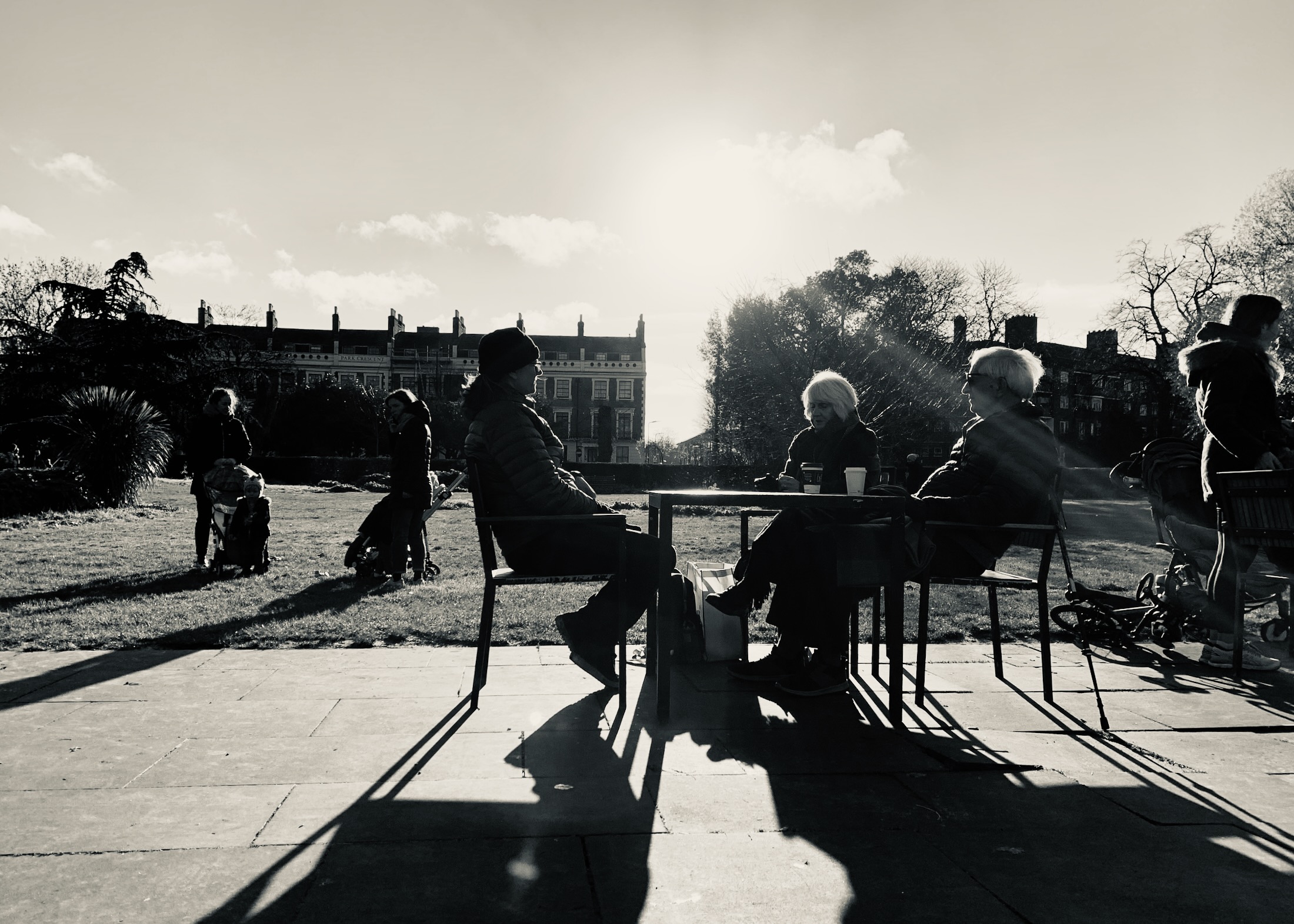 Three seated figures share a table in a sunlit park, casting long shadows while passersby stroll in the background.