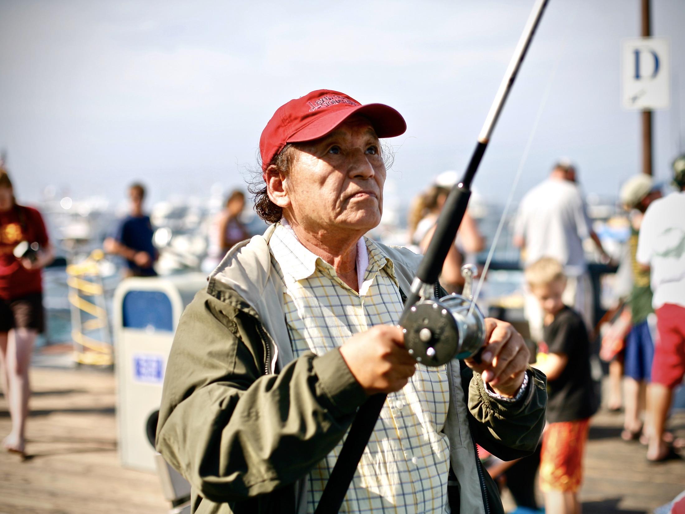 A focused fisherman in a red cap grips his fishing rod, surrounded by bustling families on a sunlit pier.
