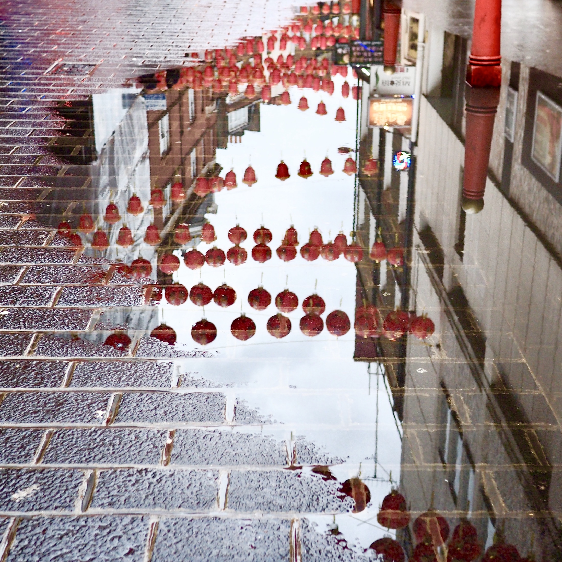 Glowing red lanterns are mirrored in a rain-soaked street, creating an enchanting urban reflection under a cloudy sky.