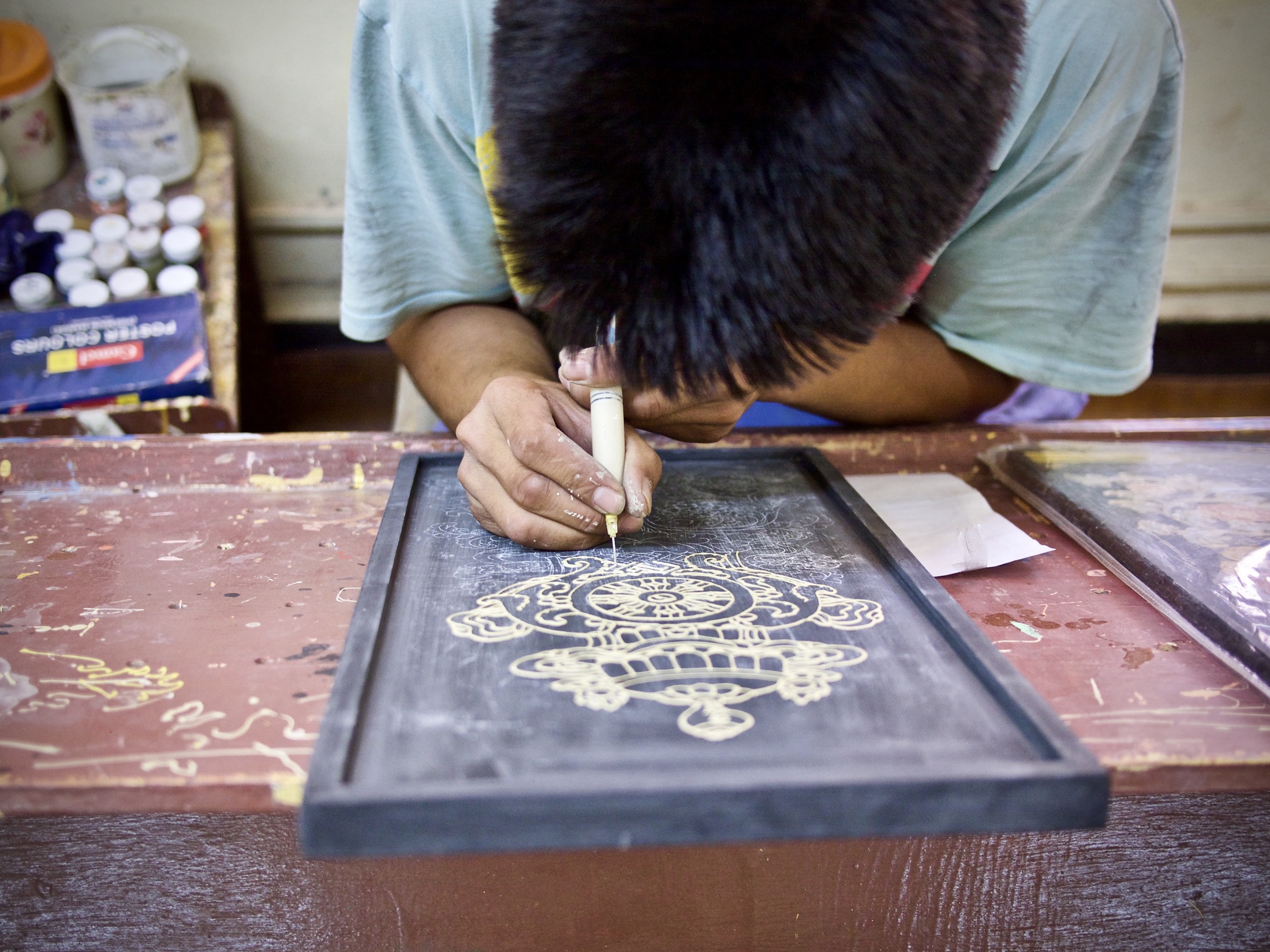 A young artist meticulously etches intricate designs onto a dark wooden panel, surrounded by vibrant paint jars.