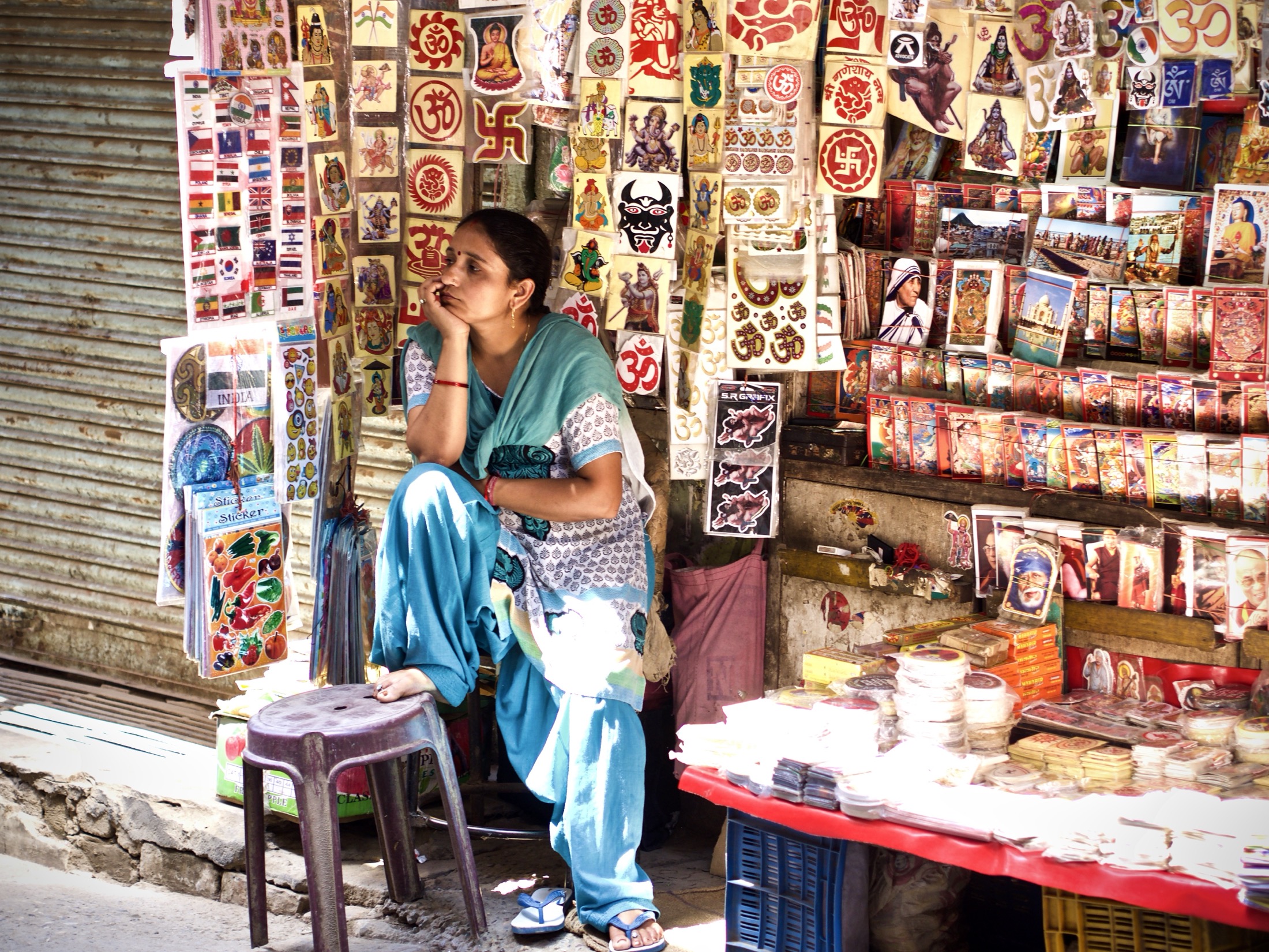 A woman sits pensively among vibrant, colorful stalls brimming with stickers and religious imagery, bathed in warm sunlight.