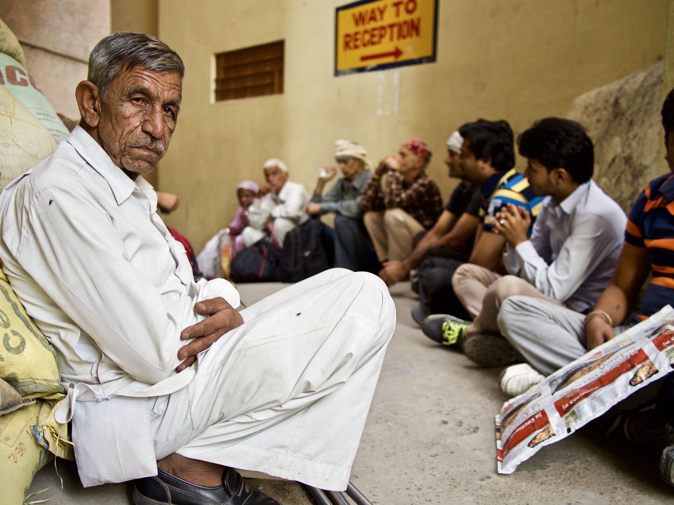 An elderly man dressed in white sits pensively against a wall while others gather in the background, creating a quiet, reflective scene.