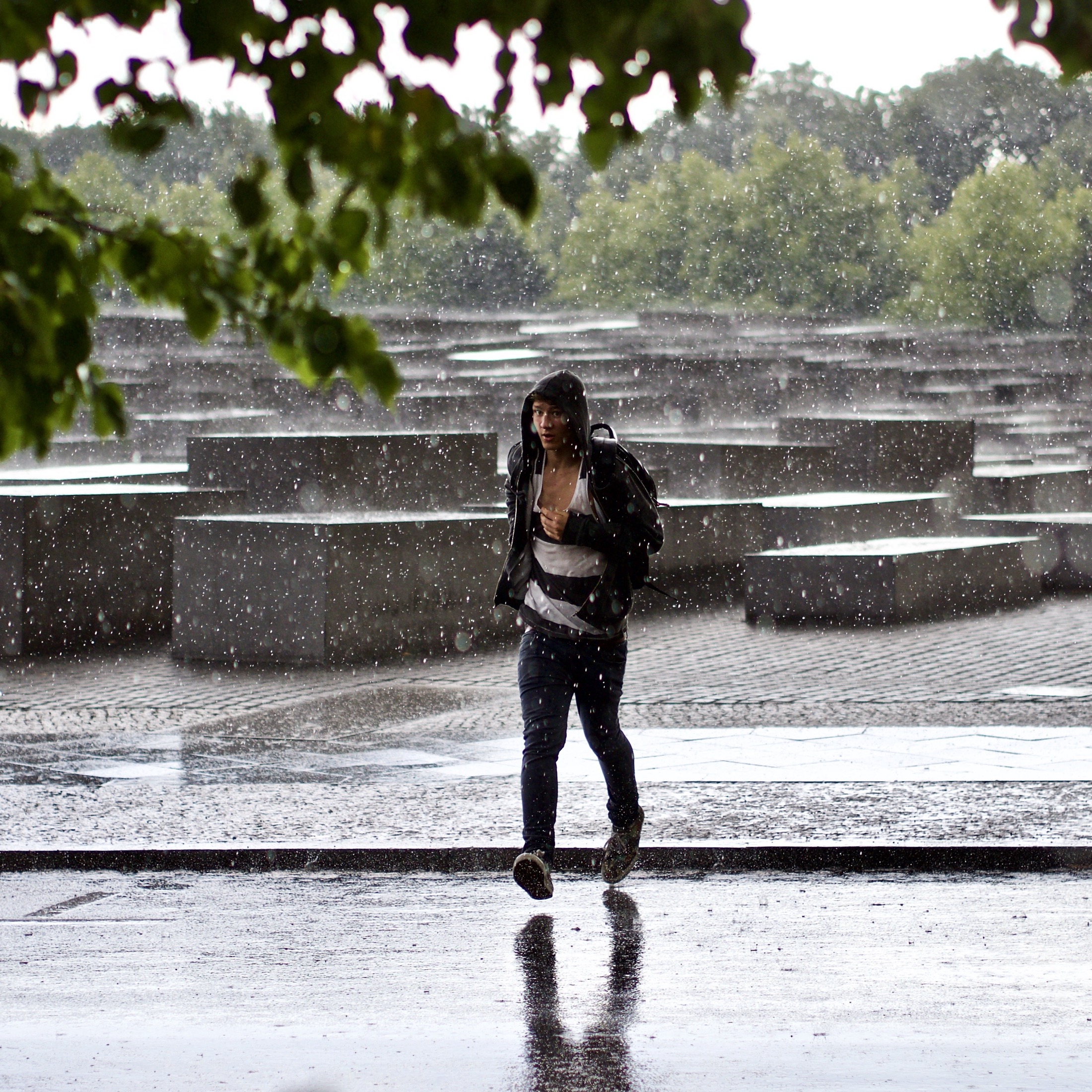 A figure strides through heavy rain, their reflection shimmering on the wet pavement amid stark, geometric monuments.