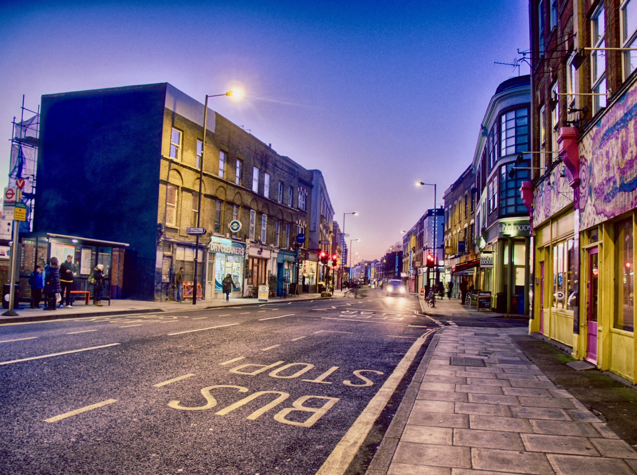 A quiet urban street at dusk, lined with colorful storefronts and a few people waiting at a bus stop under soft streetlights.